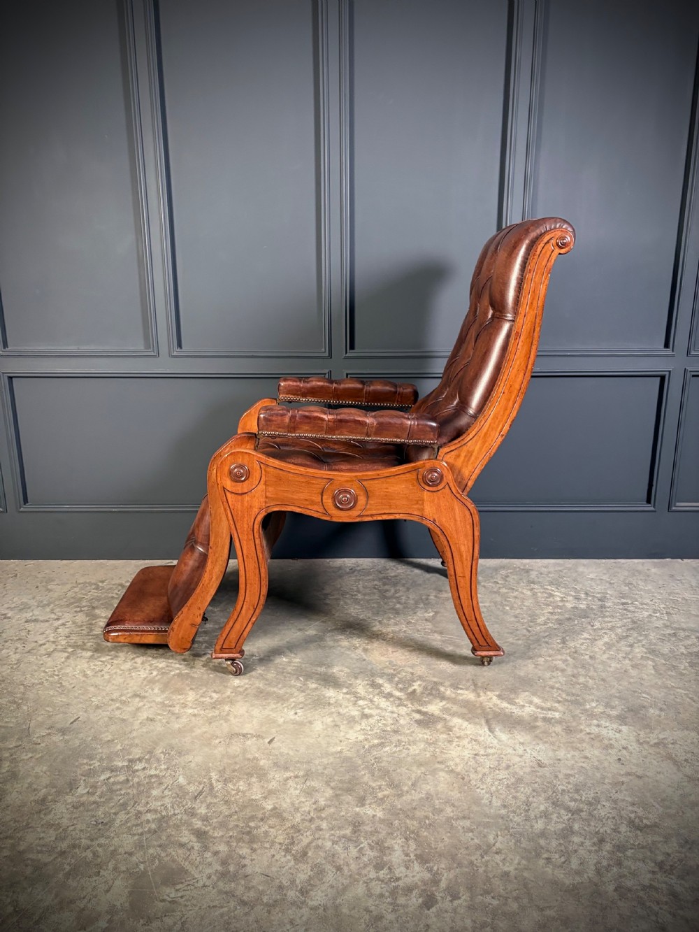 A Regency mahogany reclining armchair featuring button-tufted brown leather upholstery, padded armrests, an adjustable footrest, brass castors, and carved timber details. View IX