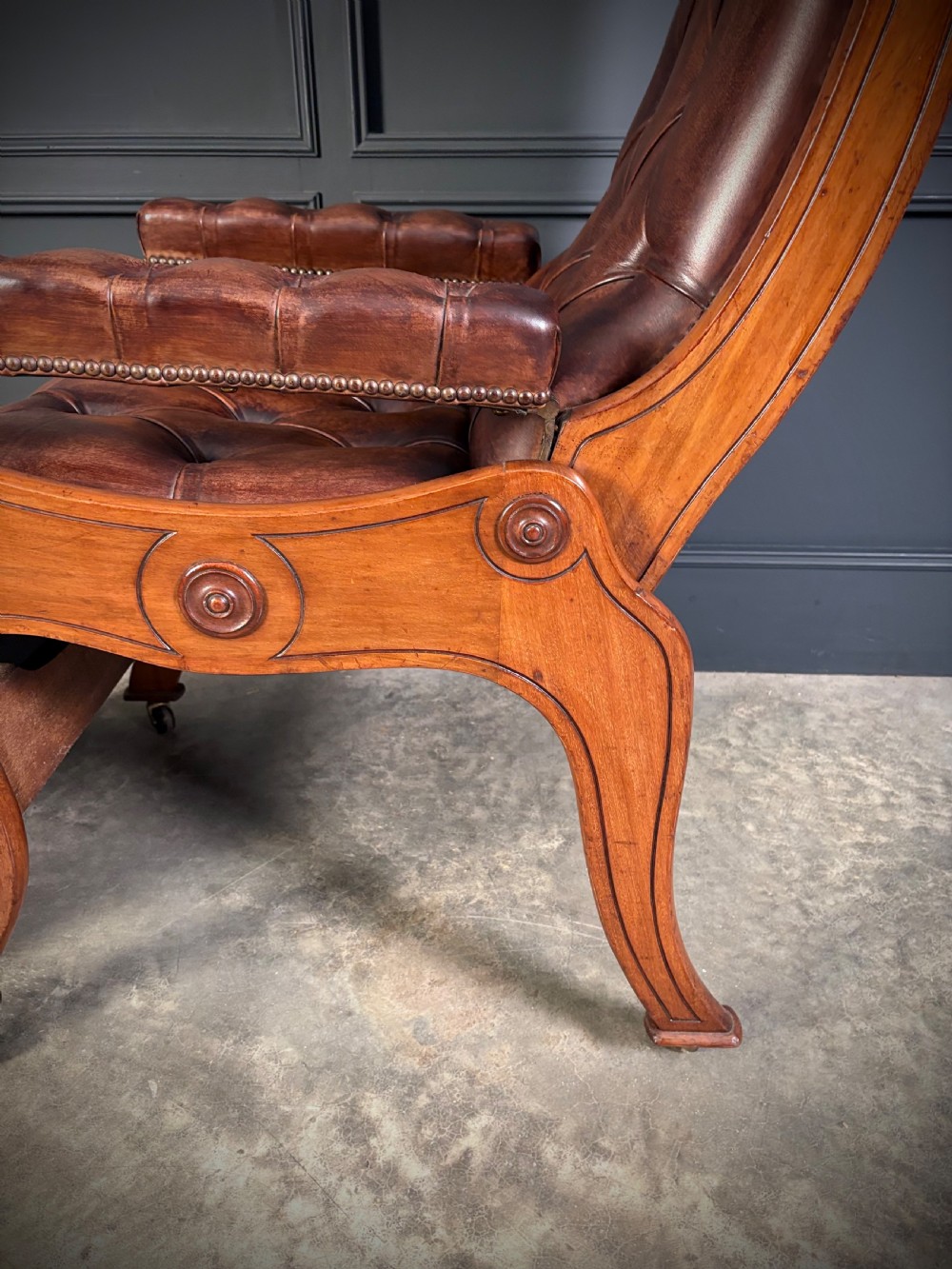A Regency mahogany reclining armchair featuring button-tufted brown leather upholstery, padded armrests, an adjustable footrest, brass castors, and carved timber details. View X
