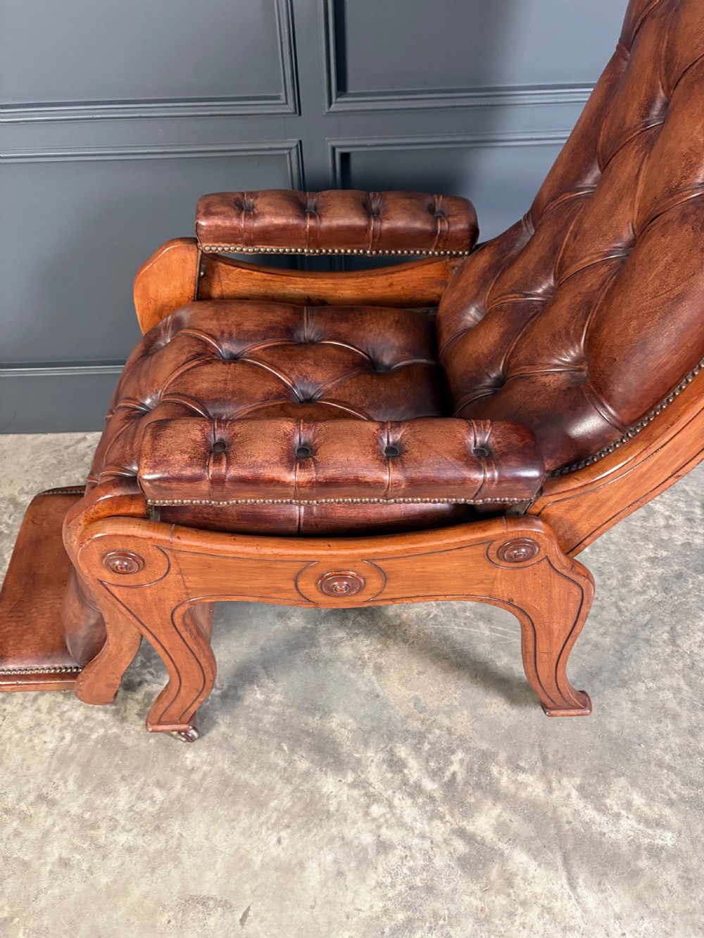 A Regency mahogany reclining armchair featuring button-tufted brown leather upholstery, padded armrests, an adjustable footrest, brass castors, and carved timber details. View XI