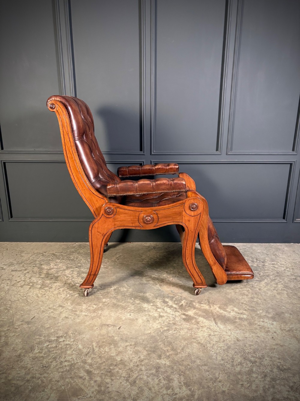 A Regency mahogany reclining armchair featuring button-tufted brown leather upholstery, padded armrests, an adjustable footrest, brass castors, and carved timber details. View XIII