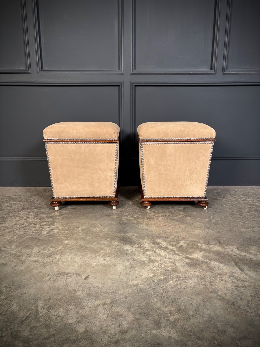 A pair of square Regency style ottomans upholstered in beige fabric with brass stud trimming, raised on short mahogany legs with castors. View II