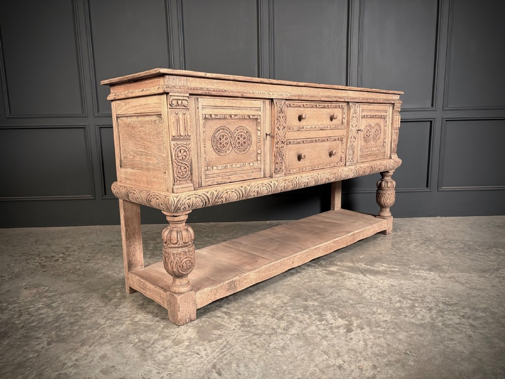 A carved bleached oak sideboard featuring geometric relief panel doors, central drawers, turned front legs, and an open lower pot board shelf.