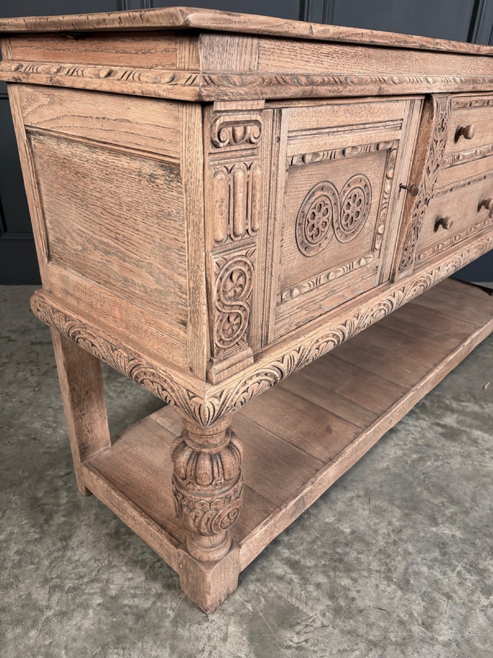 A carved bleached oak sideboard featuring geometric relief panel doors, central drawers, turned front legs, and an open lower pot board shelf. View IV