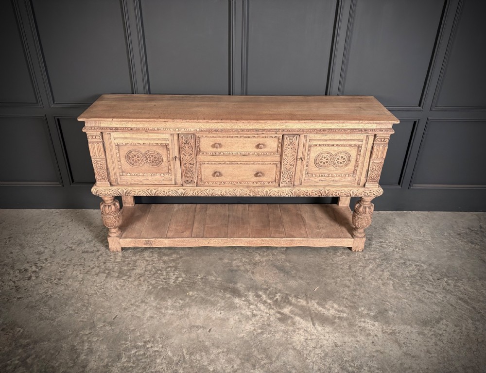 A carved bleached oak sideboard featuring geometric relief panel doors, central drawers, turned front legs, and an open lower pot board shelf. View V