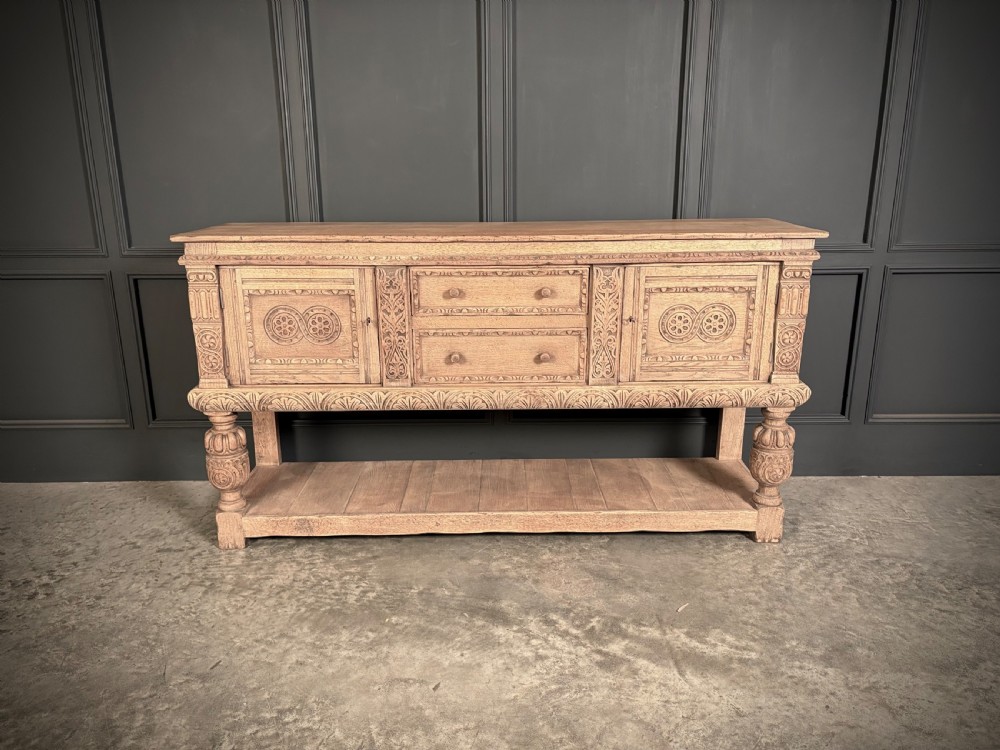 A carved bleached oak sideboard featuring geometric relief panel doors, central drawers, turned front legs, and an open lower pot board shelf. View VI