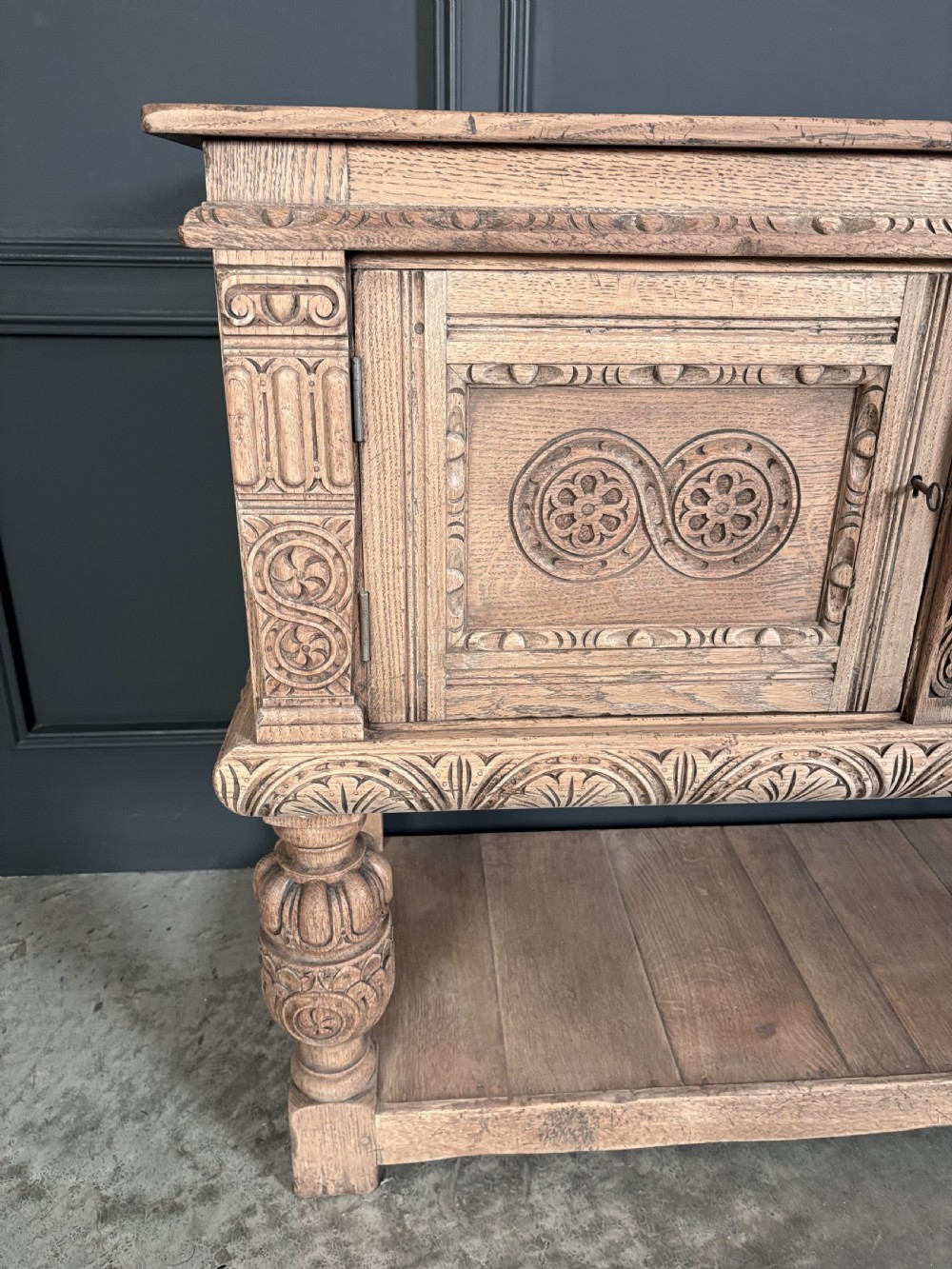 A carved bleached oak sideboard featuring geometric relief panel doors, central drawers, turned front legs, and an open lower pot board shelf. View VII