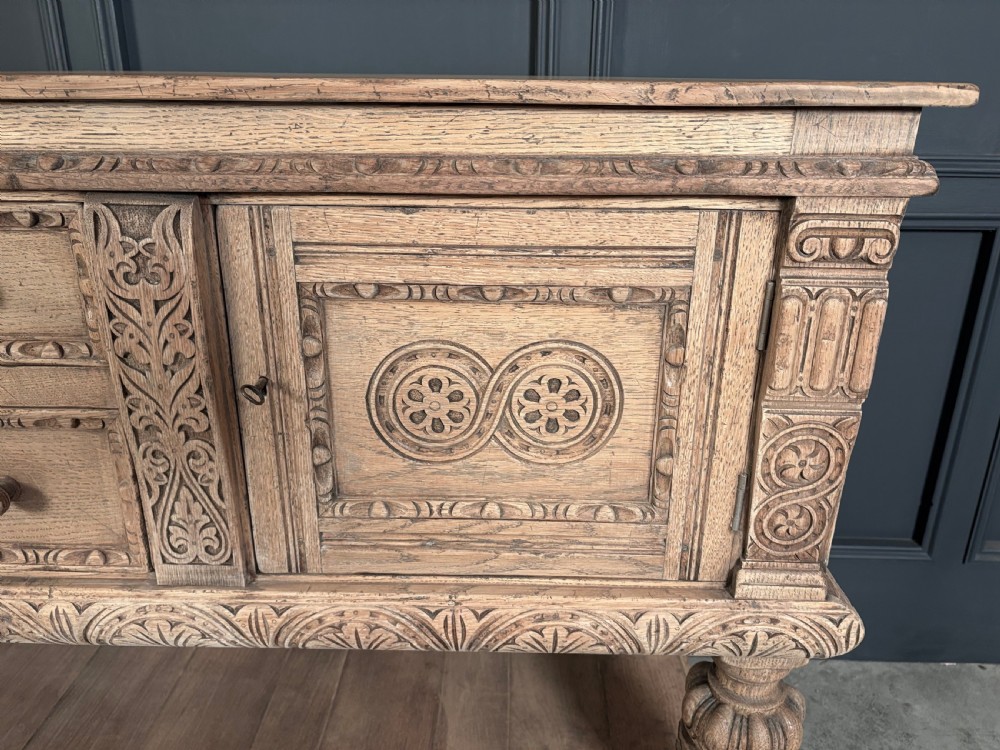 A carved bleached oak sideboard featuring geometric relief panel doors, central drawers, turned front legs, and an open lower pot board shelf. View VIII
