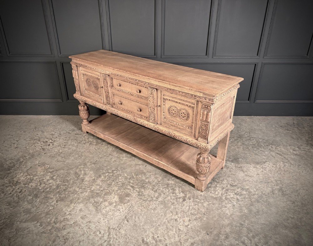 A carved bleached oak sideboard featuring geometric relief panel doors, central drawers, turned front legs, and an open lower pot board shelf. View X