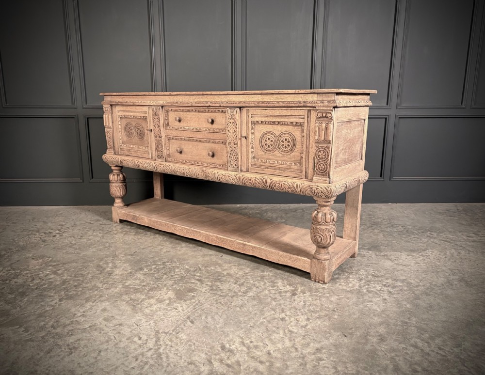 A carved bleached oak sideboard featuring geometric relief panel doors, central drawers, turned front legs, and an open lower pot board shelf. View XI