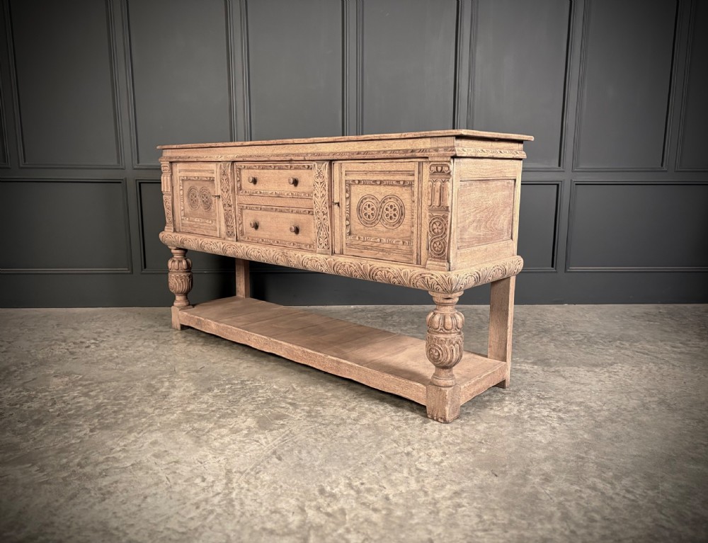 A carved bleached oak sideboard featuring geometric relief panel doors, central drawers, turned front legs, and an open lower pot board shelf. View XII