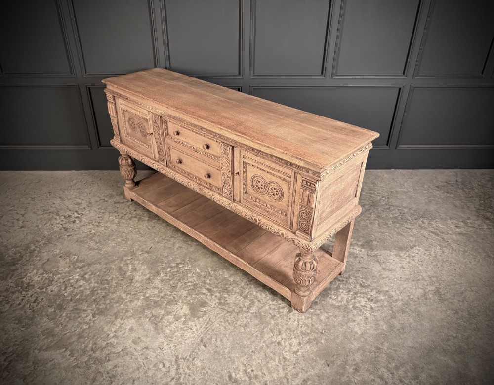 A carved bleached oak sideboard featuring geometric relief panel doors, central drawers, turned front legs, and an open lower pot board shelf. View XIII