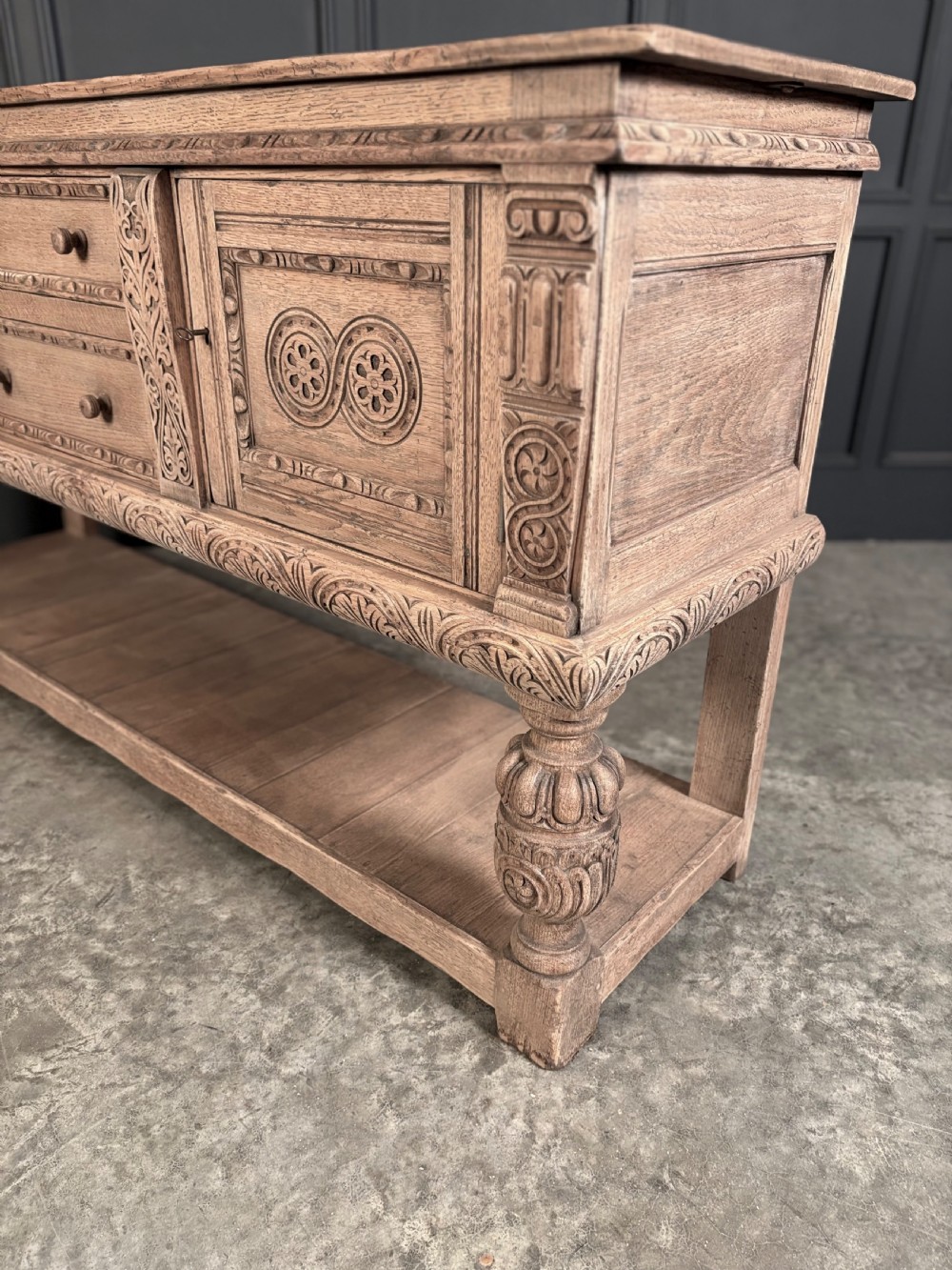 A carved bleached oak sideboard featuring geometric relief panel doors, central drawers, turned front legs, and an open lower pot board shelf. View XIV