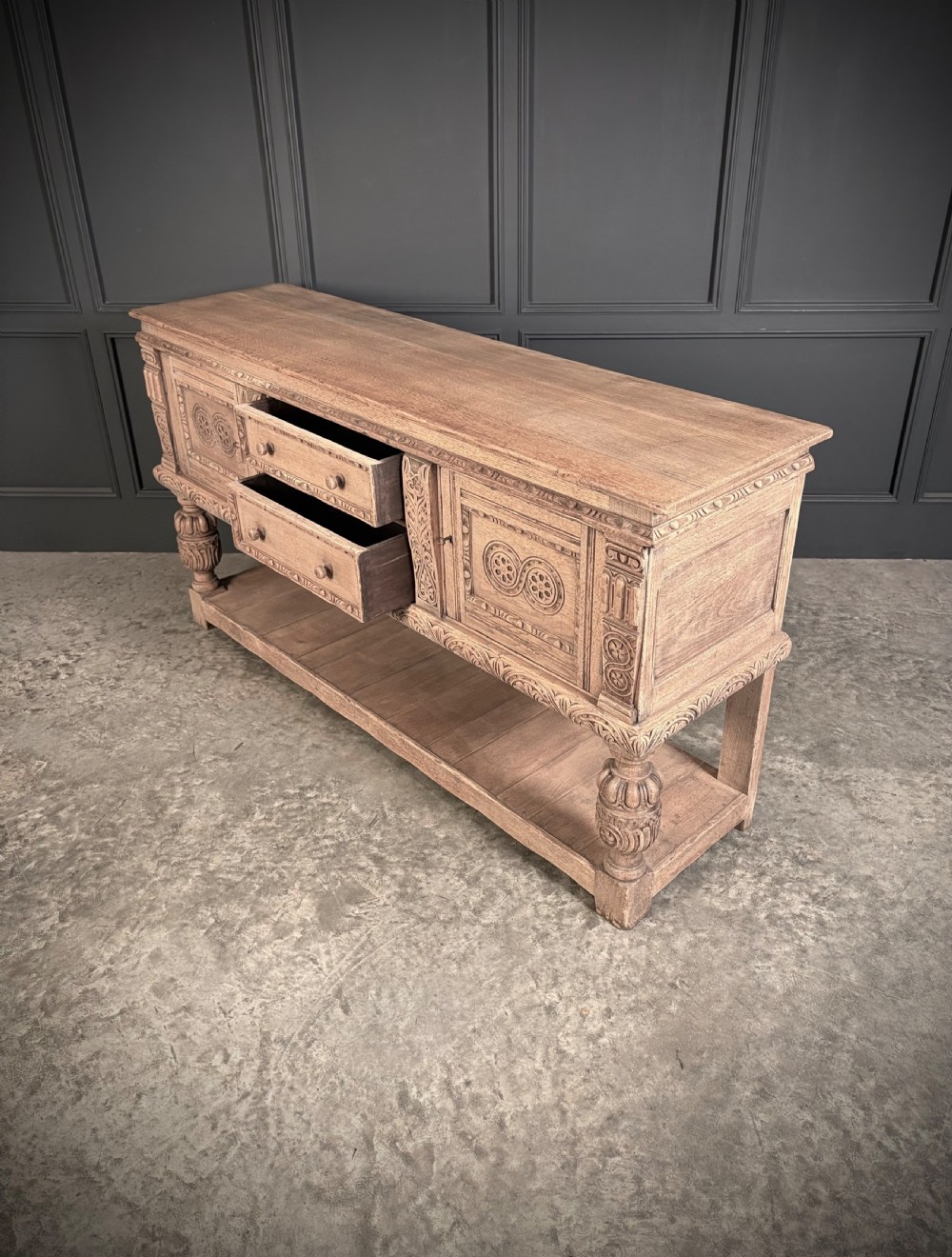 A carved bleached oak sideboard featuring geometric relief panel doors, central drawers, turned front legs, and an open lower pot board shelf. View XV