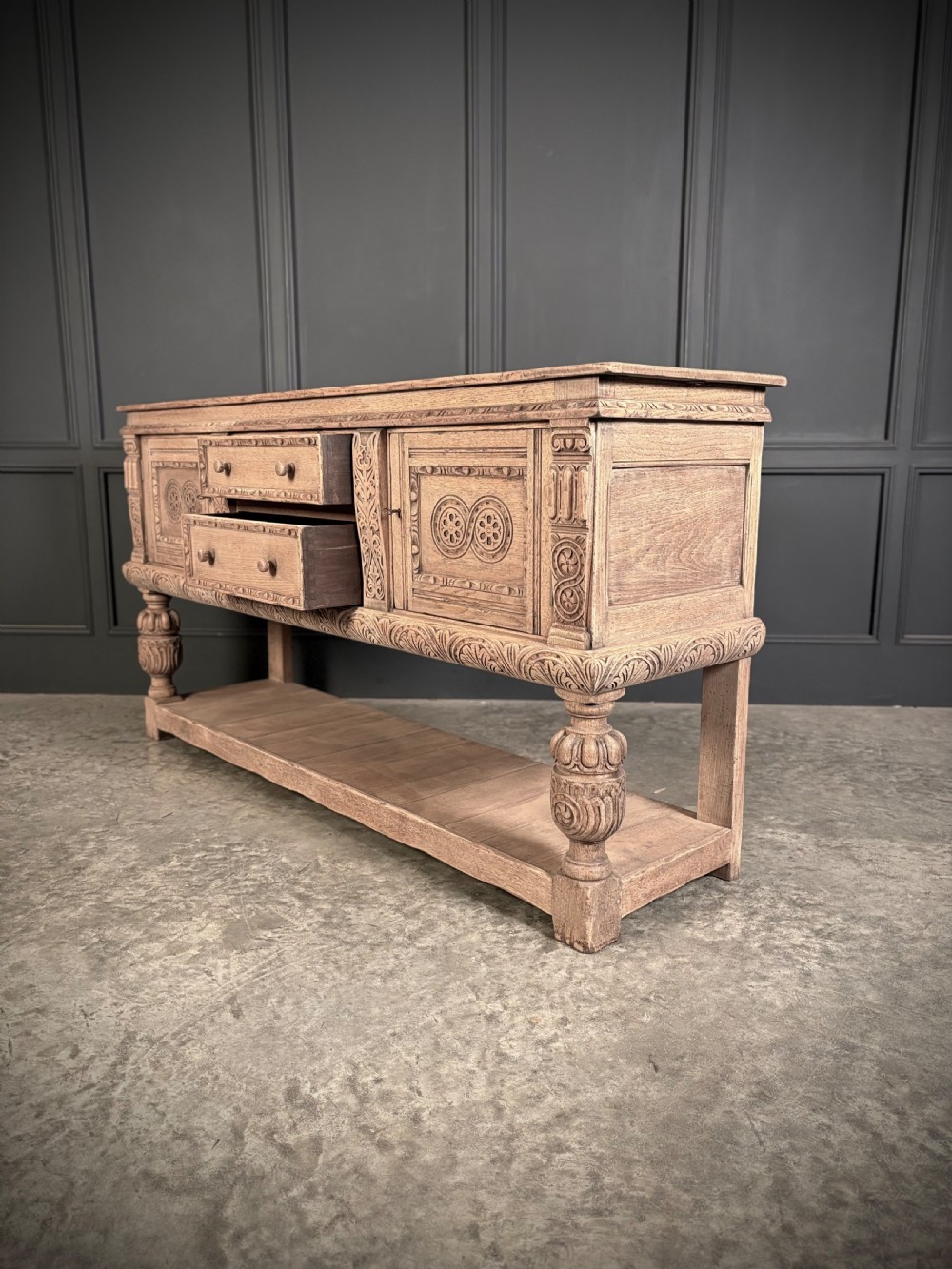 A carved bleached oak sideboard featuring geometric relief panel doors, central drawers, turned front legs, and an open lower pot board shelf. View XVI