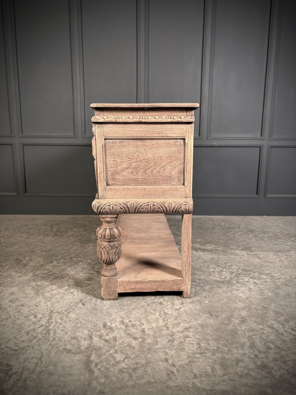 A carved bleached oak sideboard featuring geometric relief panel doors, central drawers, turned front legs, and an open lower pot board shelf. View XVII
