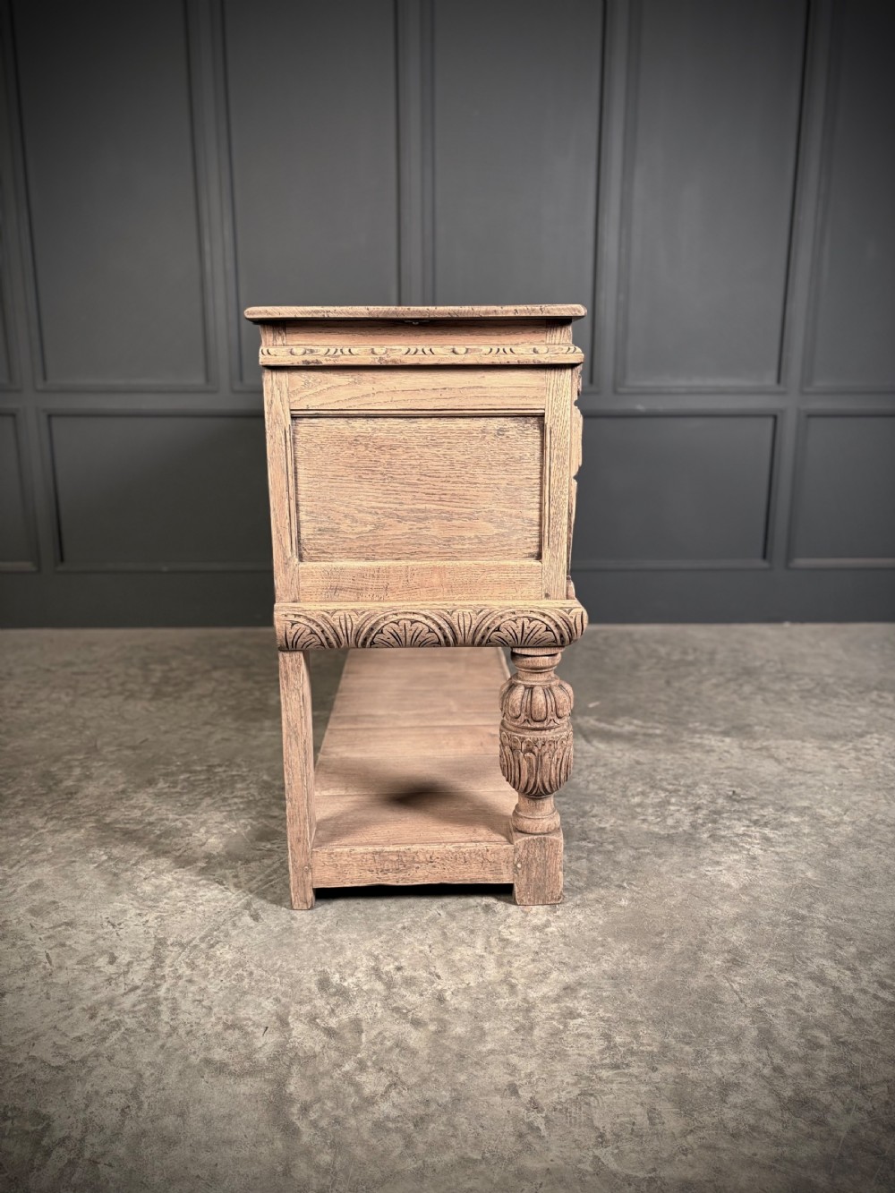 A carved bleached oak sideboard featuring geometric relief panel doors, central drawers, turned front legs, and an open lower pot board shelf. View XIX