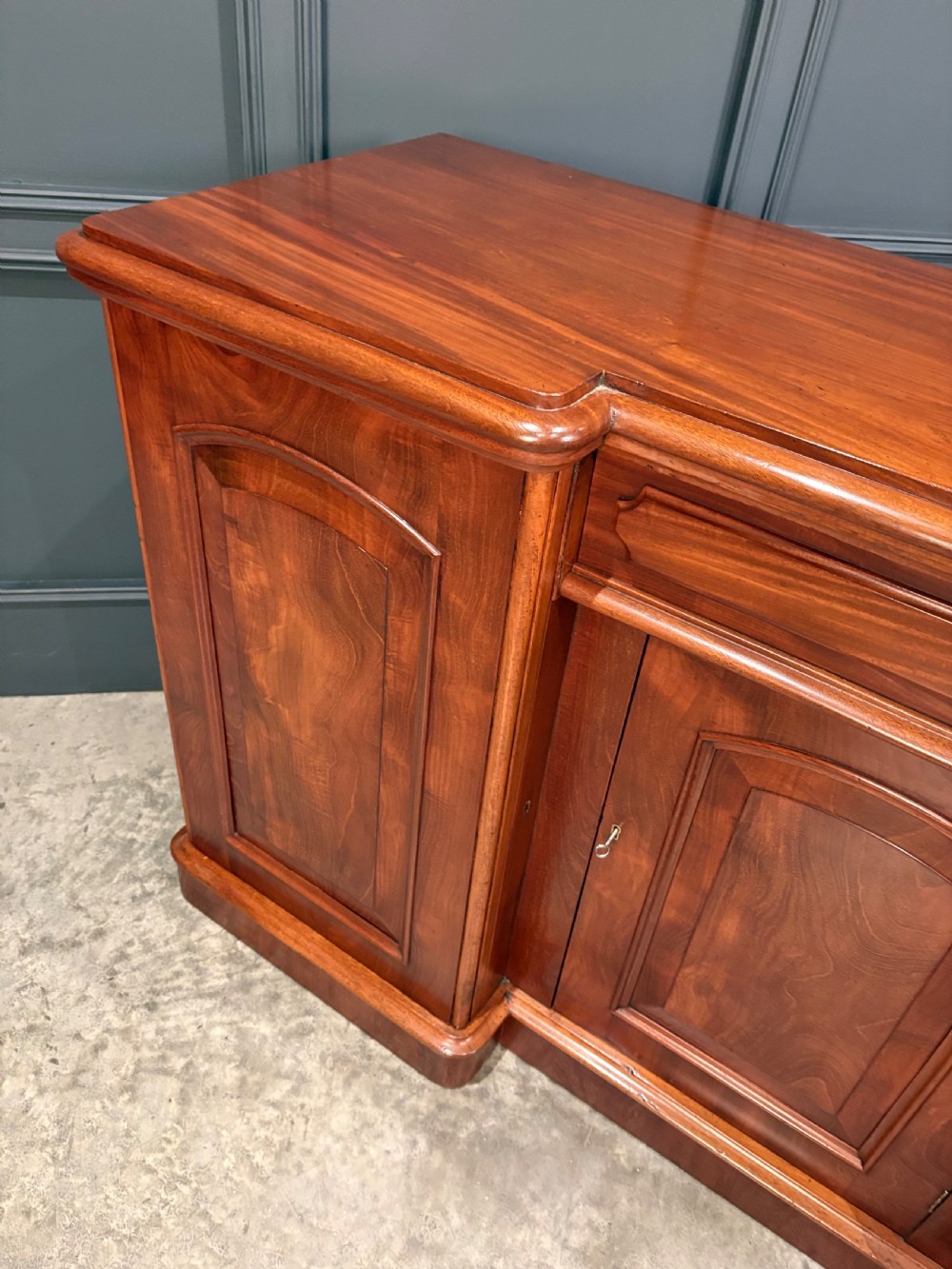 A Victorian mahogany three-door sideboard featuring flame mahogany panelled cupboard doors and a central top drawer on a solid plinth base. View XII
