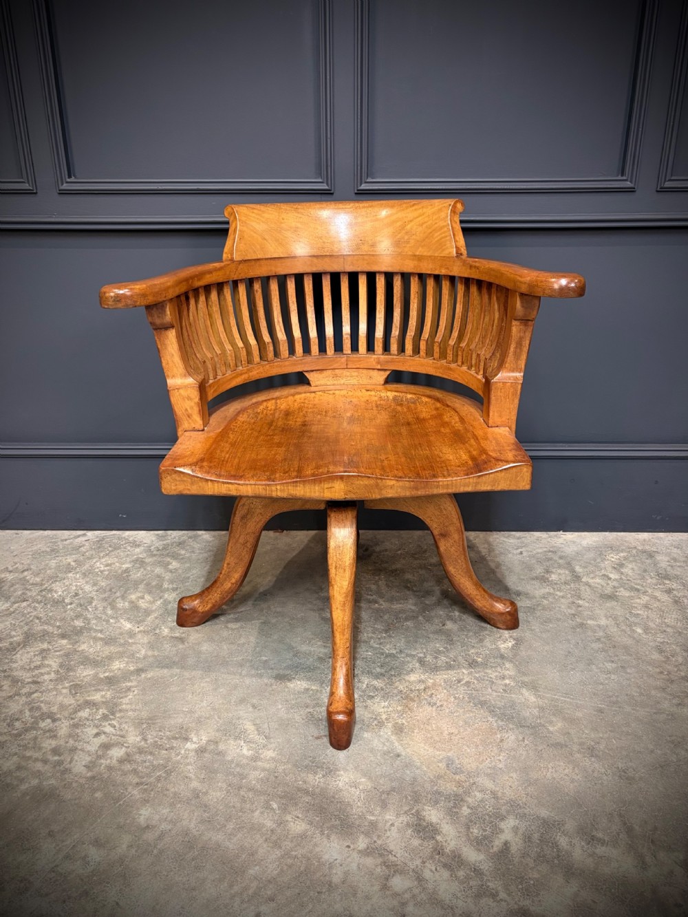 A light mahogany swivel desk chair featuring a curved slatted backrest, sweeping armrests, a shaped solid wooden seat, and a four-legged revolving base. View V