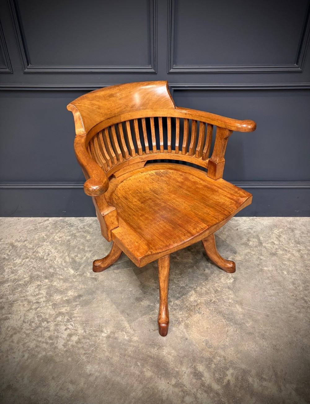 A light mahogany swivel desk chair featuring a curved slatted backrest, sweeping armrests, a shaped solid wooden seat, and a four-legged revolving base. View II