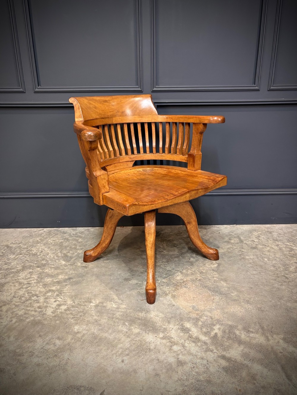 A light mahogany swivel desk chair featuring a curved slatted backrest, sweeping armrests, a shaped solid wooden seat, and a four-legged revolving base. View III