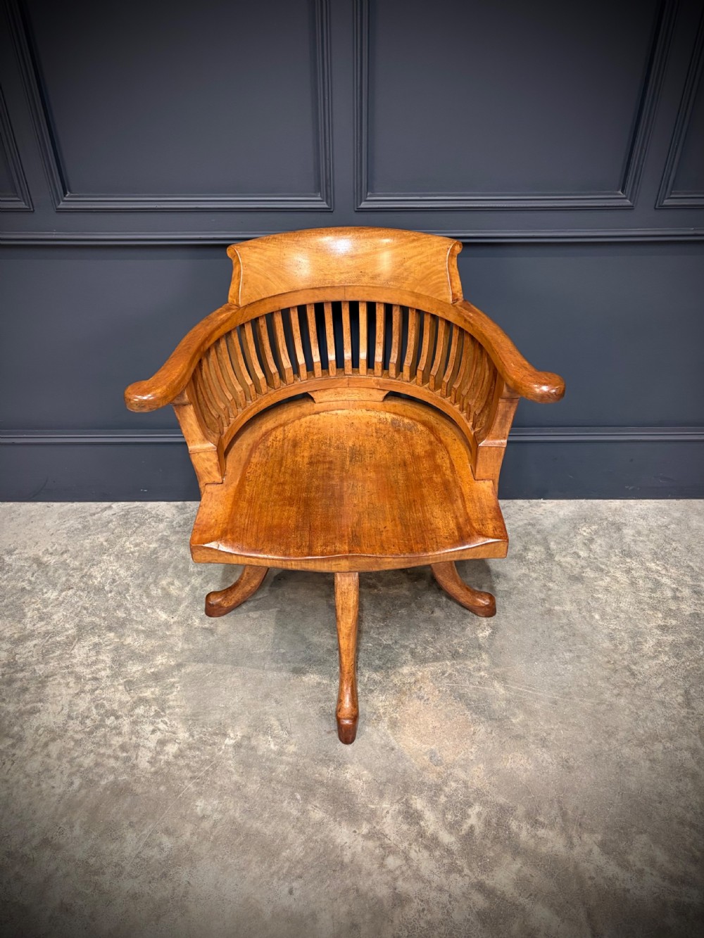 A light mahogany swivel desk chair featuring a curved slatted backrest, sweeping armrests, a shaped solid wooden seat, and a four-legged revolving base. View IV