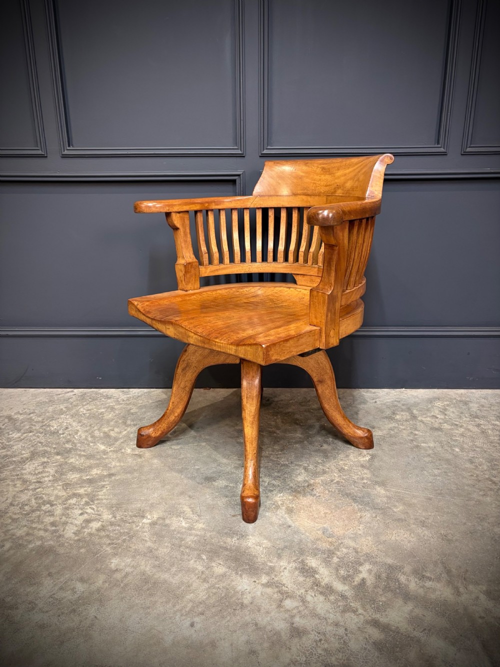 A light mahogany swivel desk chair featuring a curved slatted backrest, sweeping armrests, a shaped solid wooden seat, and a four-legged revolving base.