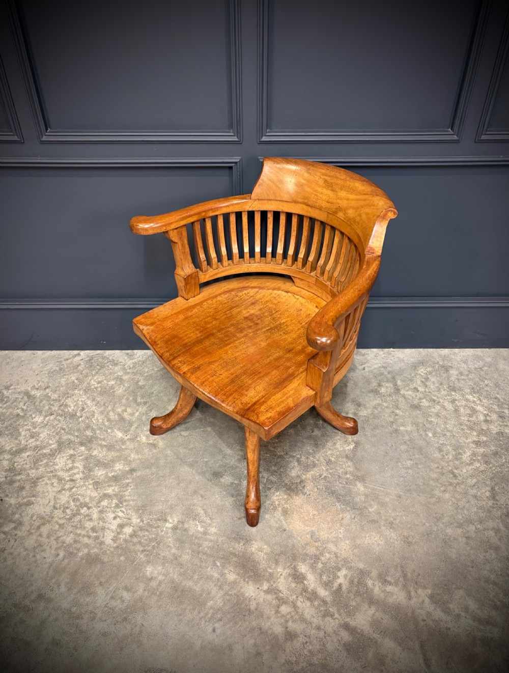 A light mahogany swivel desk chair featuring a curved slatted backrest, sweeping armrests, a shaped solid wooden seat, and a four-legged revolving base. View VI