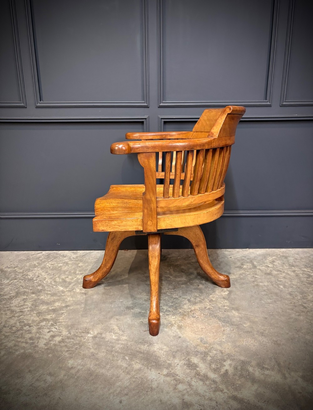 A light mahogany swivel desk chair featuring a curved slatted backrest, sweeping armrests, a shaped solid wooden seat, and a four-legged revolving base. View VII