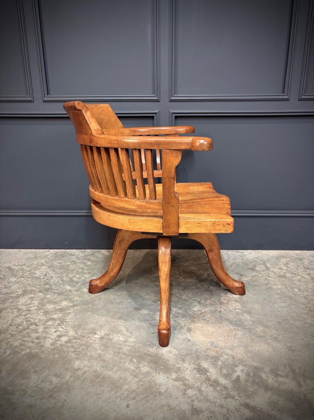 A light mahogany swivel desk chair featuring a curved slatted backrest, sweeping armrests, a shaped solid wooden seat, and a four-legged revolving base. View IX