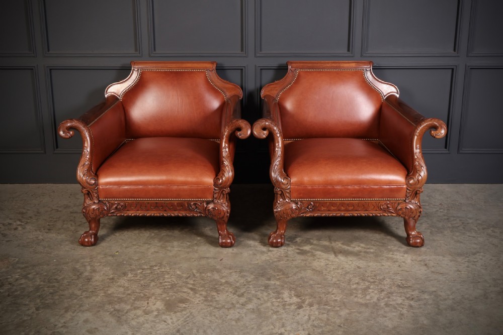 A pair of carved mahogany armchairs upholstered in warm chestnut brown leather with brass stud detailing and ornate scroll arms.