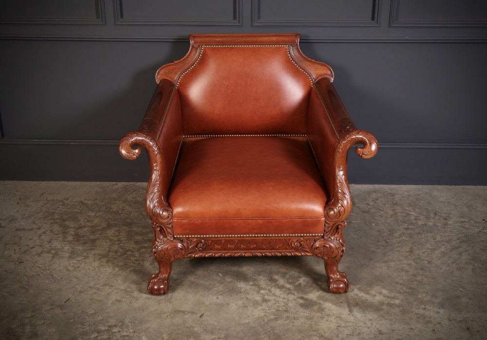 A pair of carved mahogany armchairs upholstered in warm chestnut brown leather with brass stud detailing and ornate scroll arms. View V