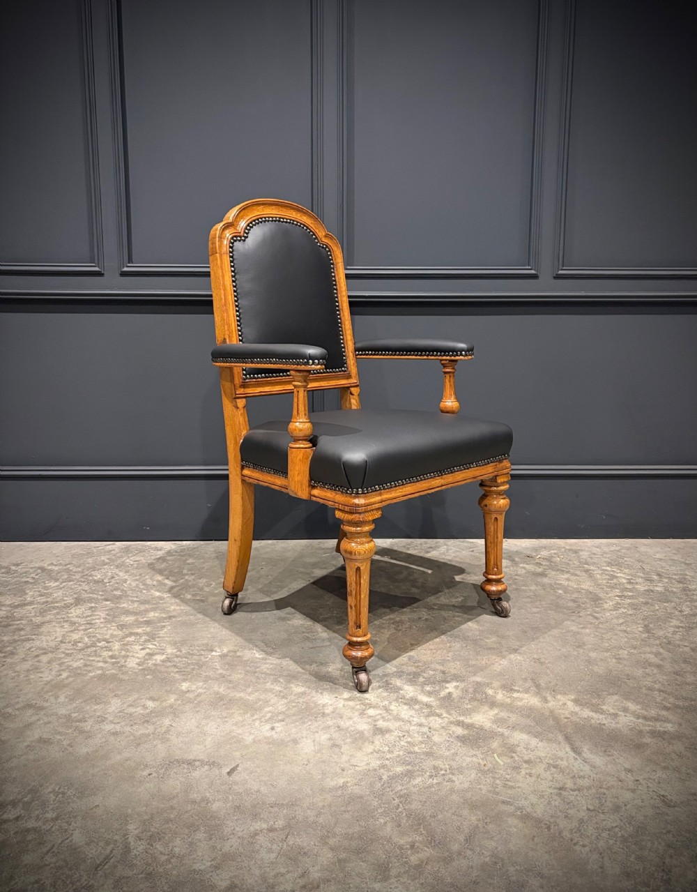 Victorian solid oak armchair featuring black leather upholstery with studding, padded armrests, turned front legs, and brass castors.