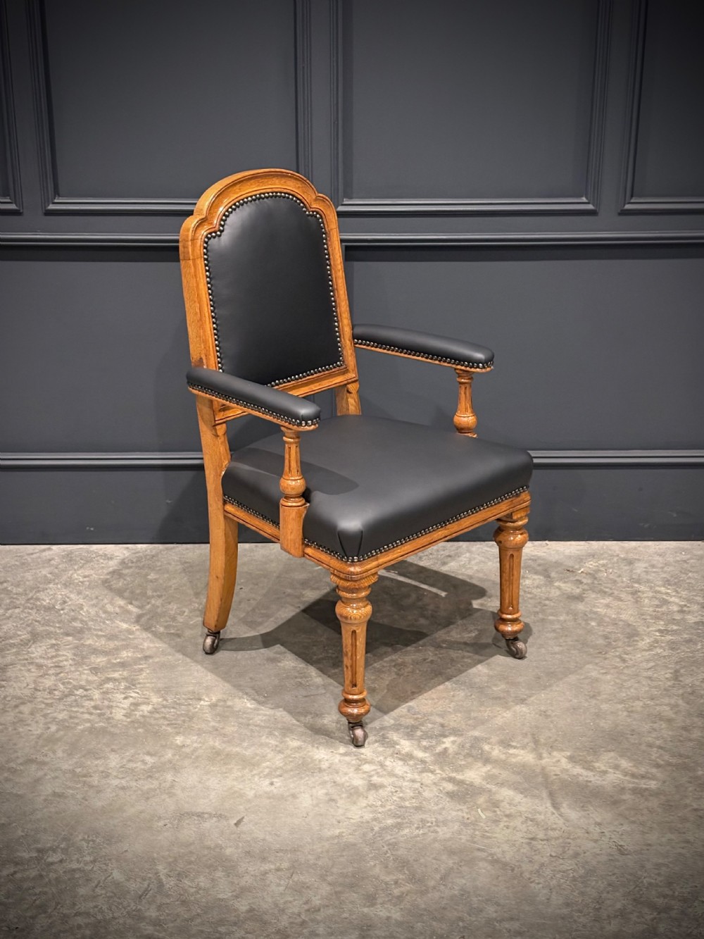 Victorian solid oak armchair featuring black leather upholstery with studding, padded armrests, turned front legs, and brass castors. View II