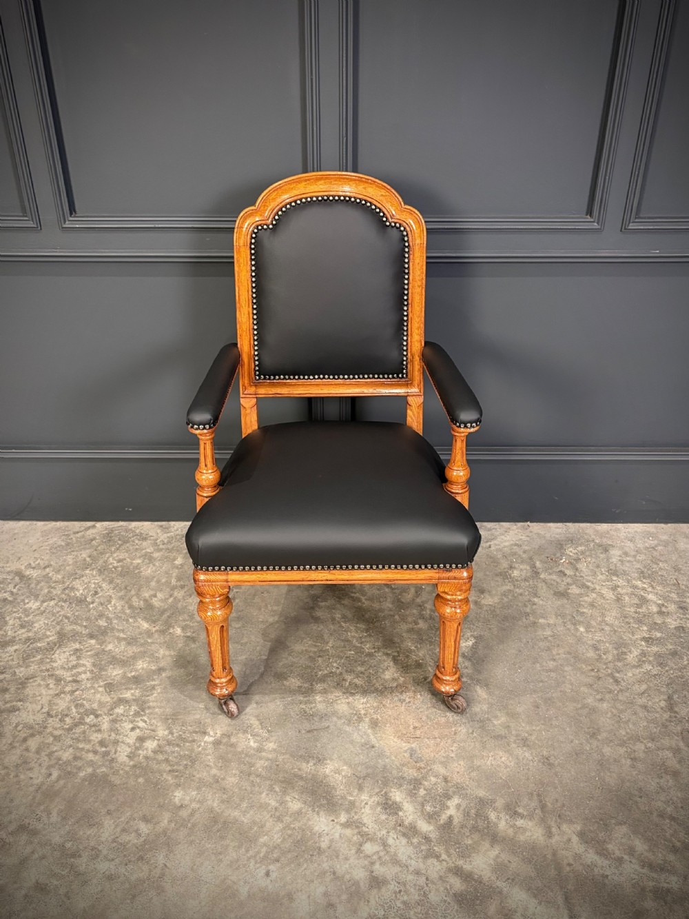 Victorian solid oak armchair featuring black leather upholstery with studding, padded armrests, turned front legs, and brass castors. View III