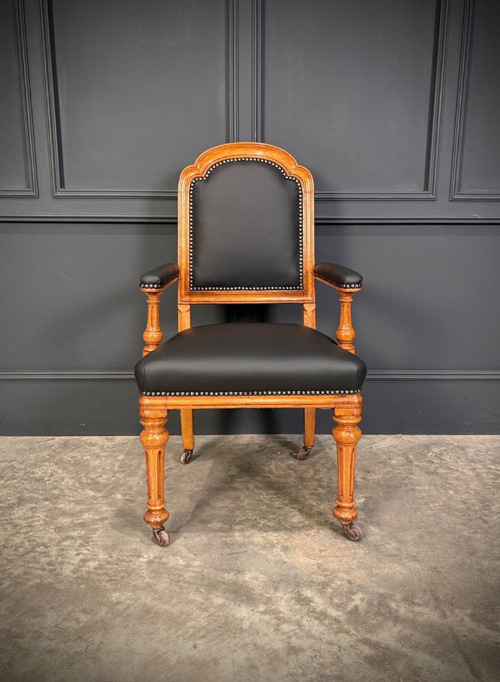 Victorian solid oak armchair featuring black leather upholstery with studding, padded armrests, turned front legs, and brass castors. View IV