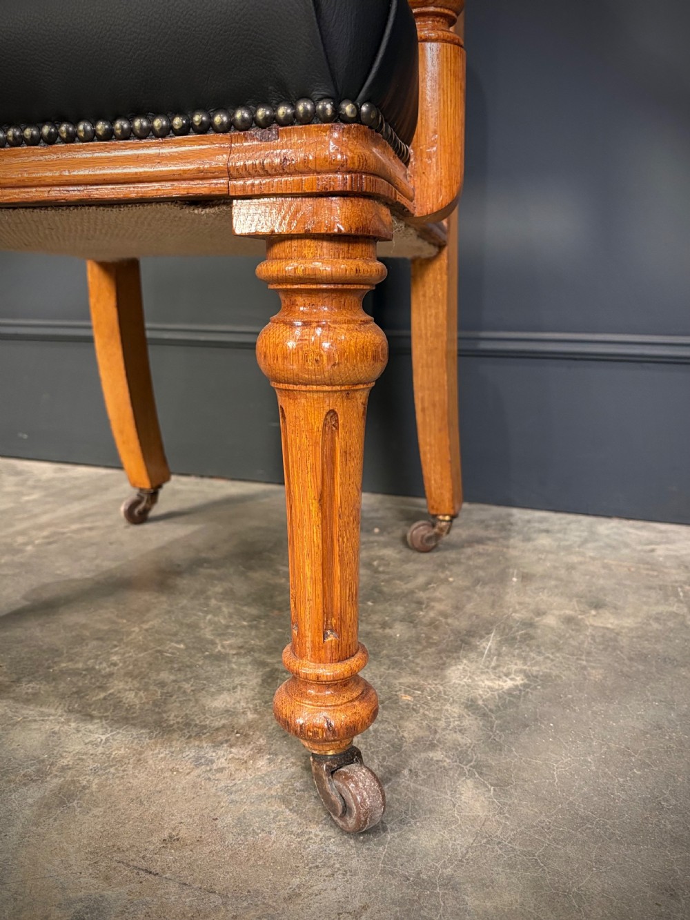 Victorian solid oak armchair featuring black leather upholstery with studding, padded armrests, turned front legs, and brass castors. View V