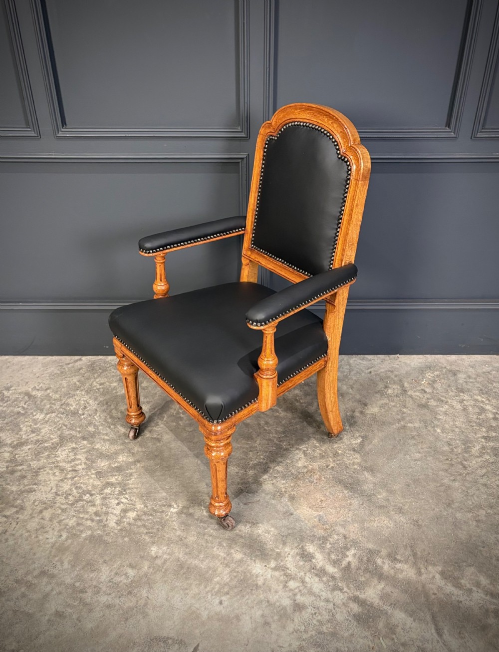 Victorian solid oak armchair featuring black leather upholstery with studding, padded armrests, turned front legs, and brass castors. View VI