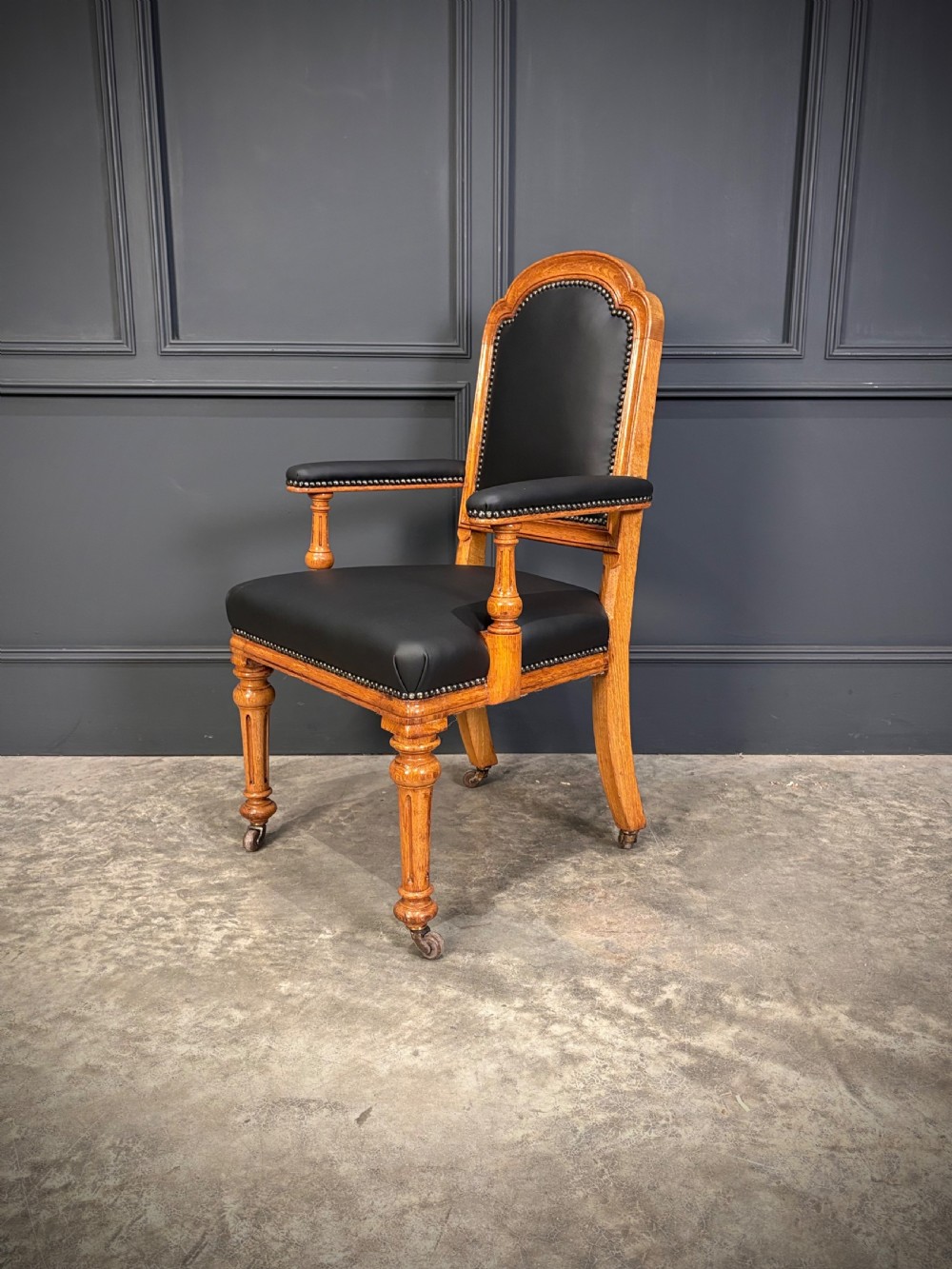 Victorian solid oak armchair featuring black leather upholstery with studding, padded armrests, turned front legs, and brass castors. View VII