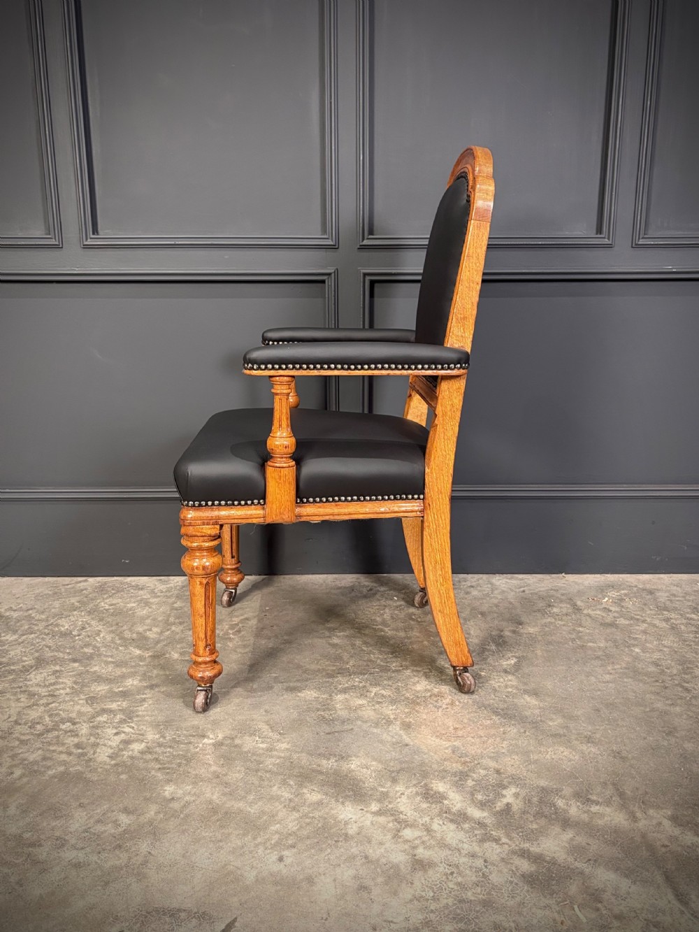 Victorian solid oak armchair featuring black leather upholstery with studding, padded armrests, turned front legs, and brass castors. View VIII