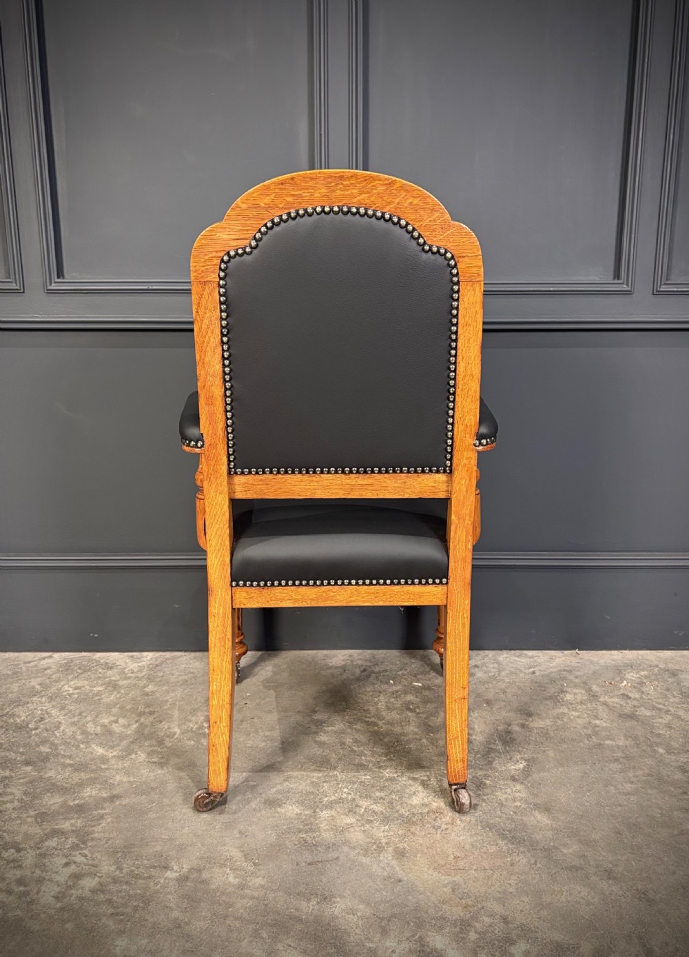 Victorian solid oak armchair featuring black leather upholstery with studding, padded armrests, turned front legs, and brass castors. View IX
