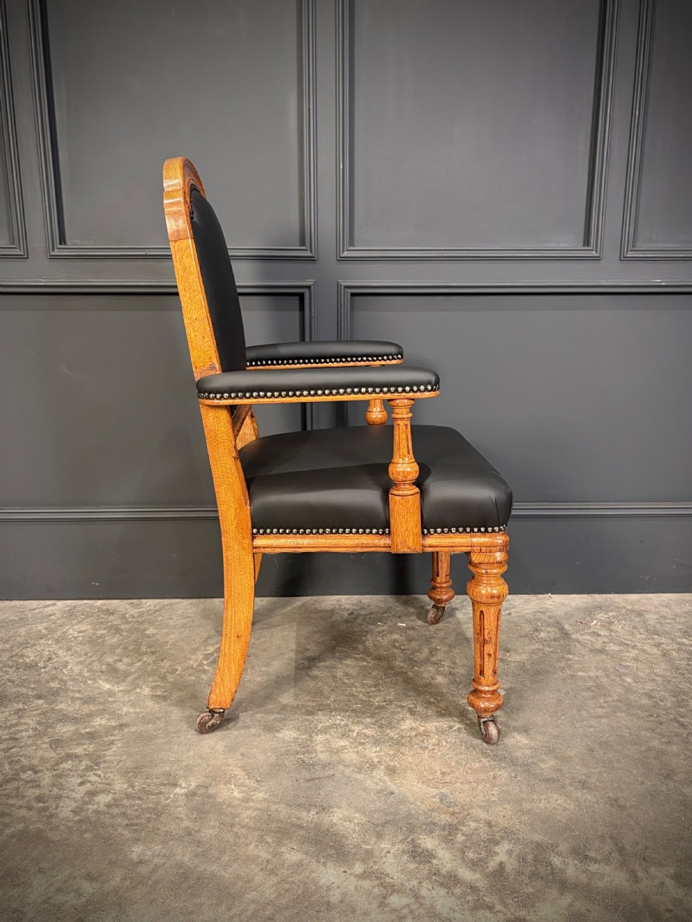 Victorian solid oak armchair featuring black leather upholstery with studding, padded armrests, turned front legs, and brass castors. View X