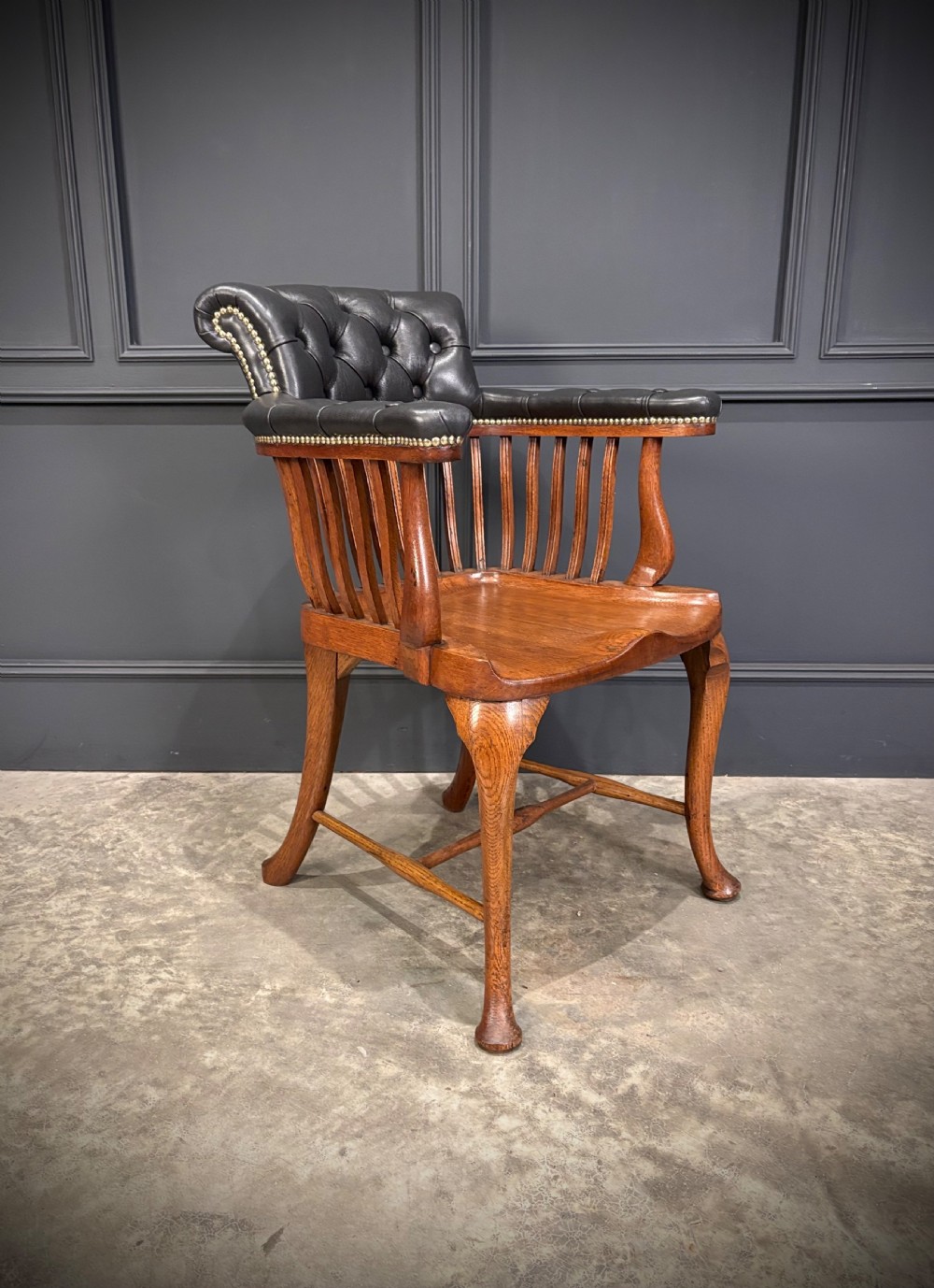 A Victorian oak desk chair featuring a deep-buttoned black leather top rail with brass studs, an open spindle back, shaped wooden seat, and cabriole front legs.