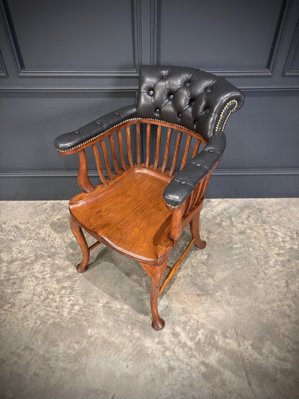 A Victorian oak desk chair featuring a deep-buttoned black leather top rail with brass studs, an open spindle back, shaped wooden seat, and cabriole front legs. View VI