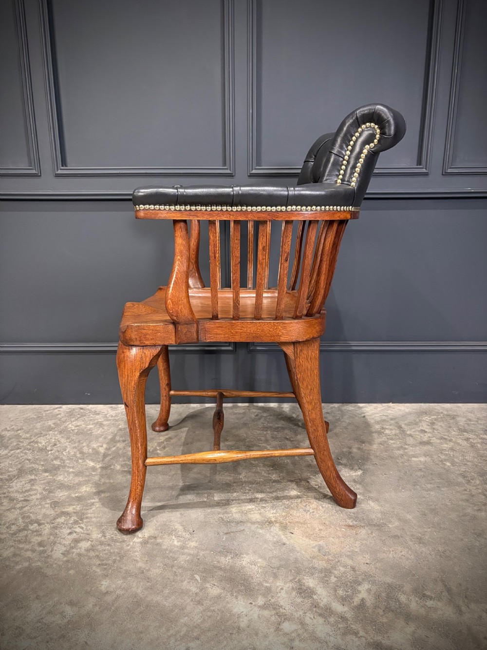 A Victorian oak desk chair featuring a deep-buttoned black leather top rail with brass studs, an open spindle back, shaped wooden seat, and cabriole front legs. View VII