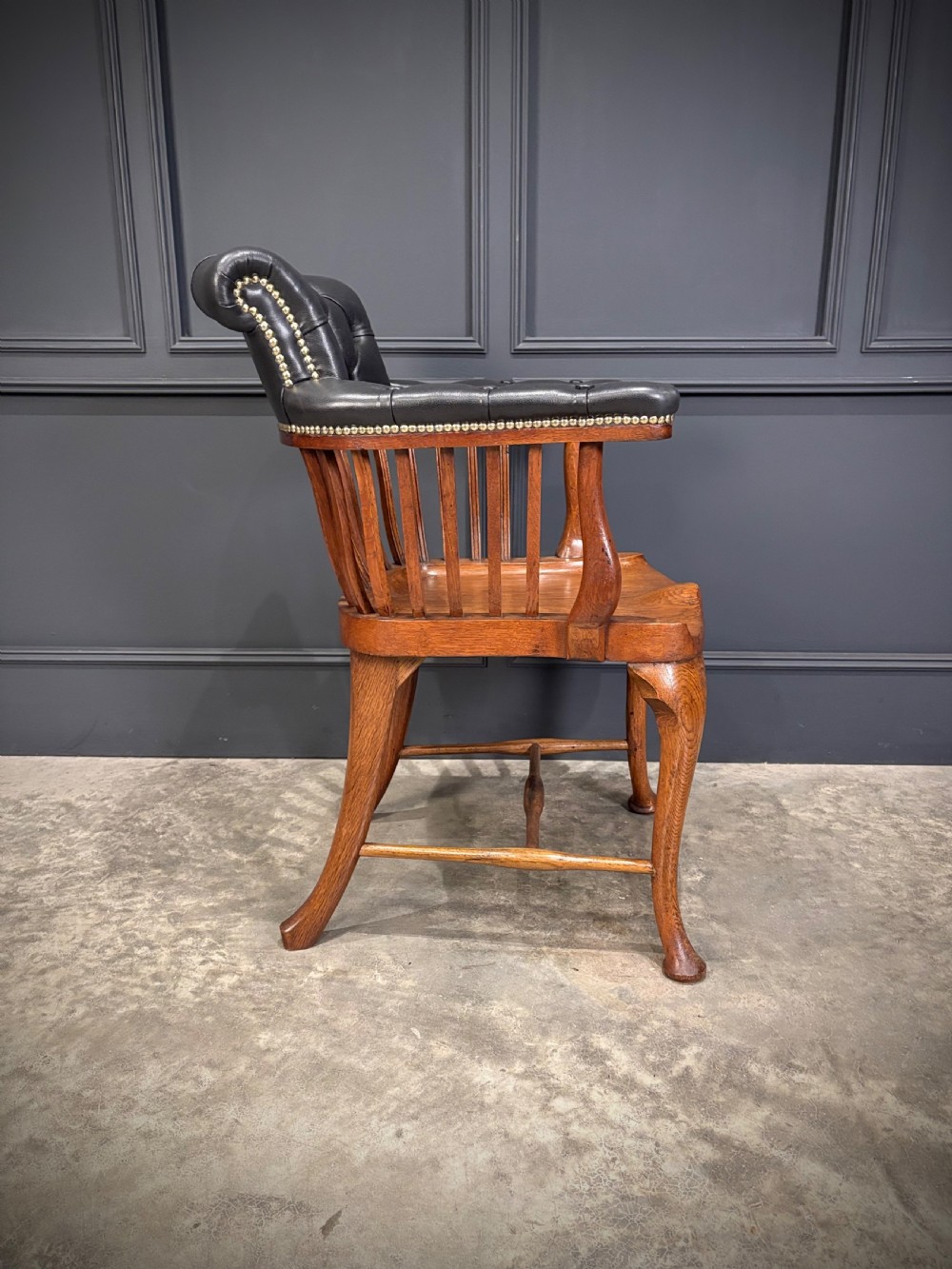 A Victorian oak desk chair featuring a deep-buttoned black leather top rail with brass studs, an open spindle back, shaped wooden seat, and cabriole front legs. View IX
