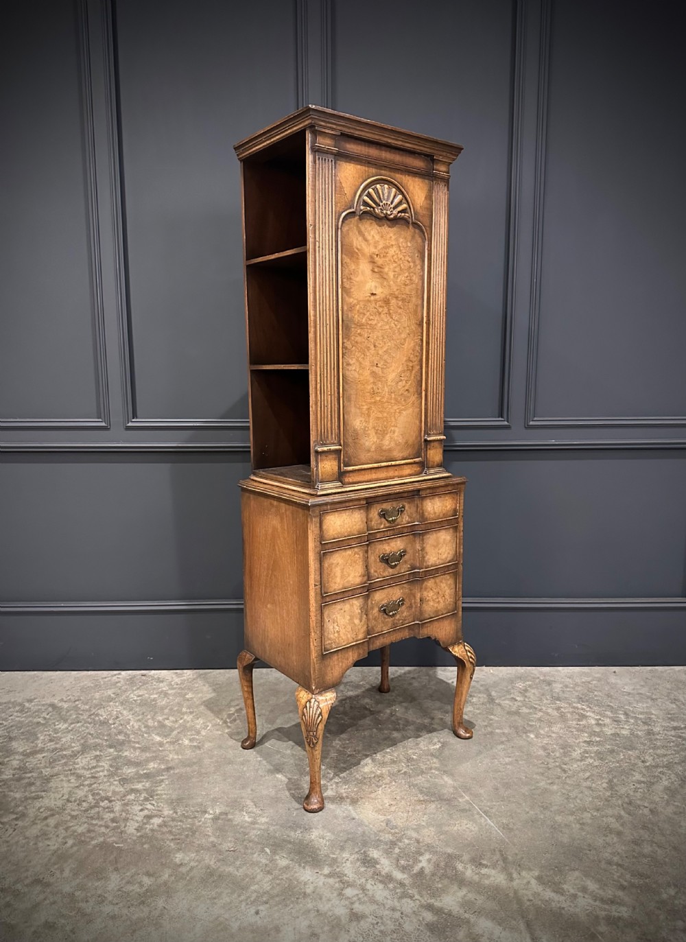 A tall, narrow Queen Anne style walnut bookcase featuring an arched burr walnut front panel, open side shelving, three drawers with brass handles, and cabriole legs with carved shell knees.