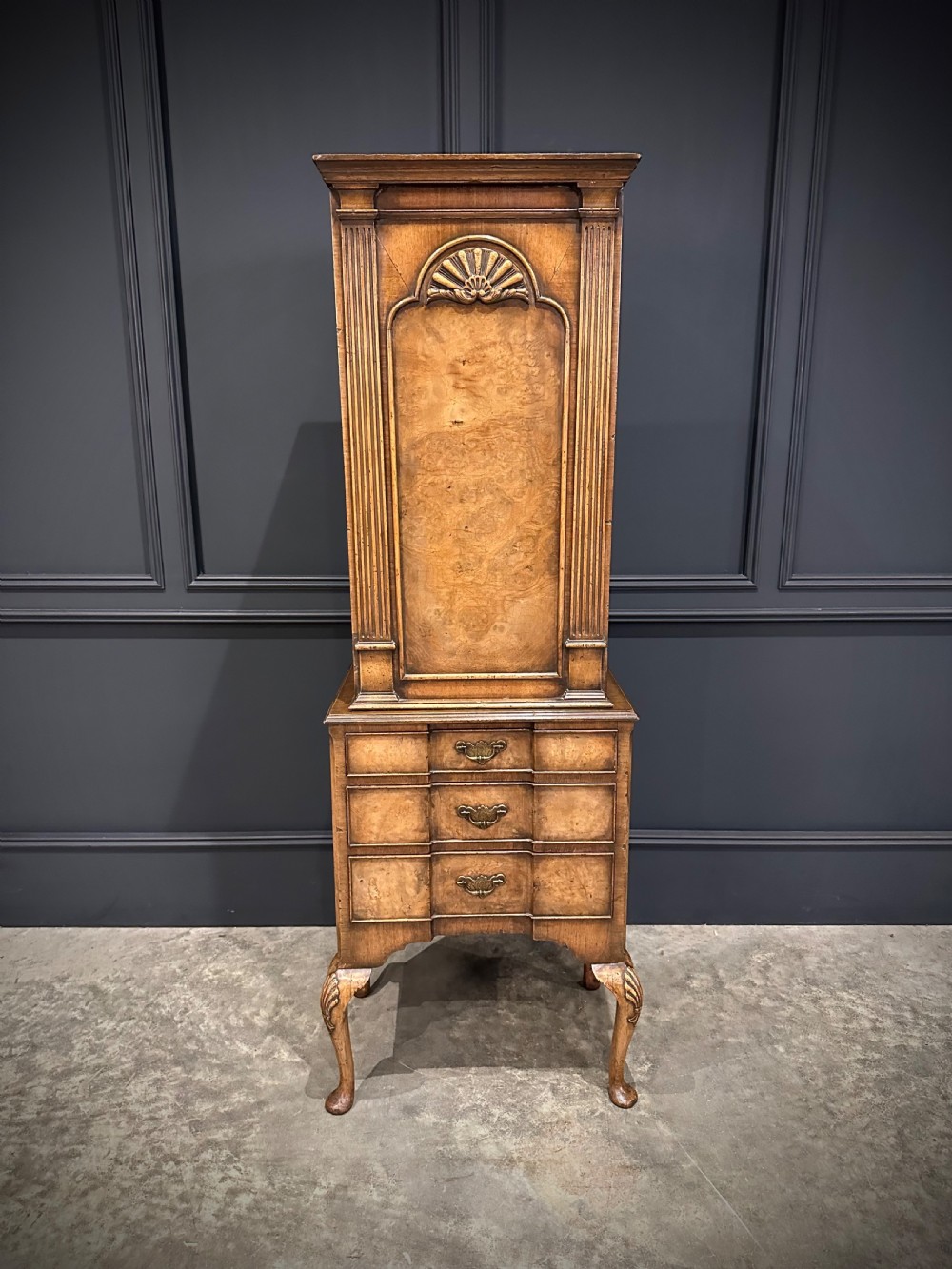 A tall, narrow Queen Anne style walnut bookcase featuring an arched burr walnut front panel, open side shelving, three drawers with brass handles, and cabriole legs with carved shell knees. View II