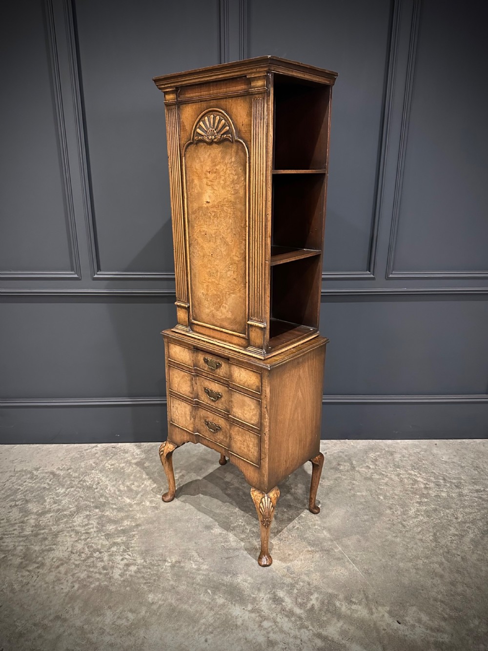 A tall, narrow Queen Anne style walnut bookcase featuring an arched burr walnut front panel, open side shelving, three drawers with brass handles, and cabriole legs with carved shell knees. View VI