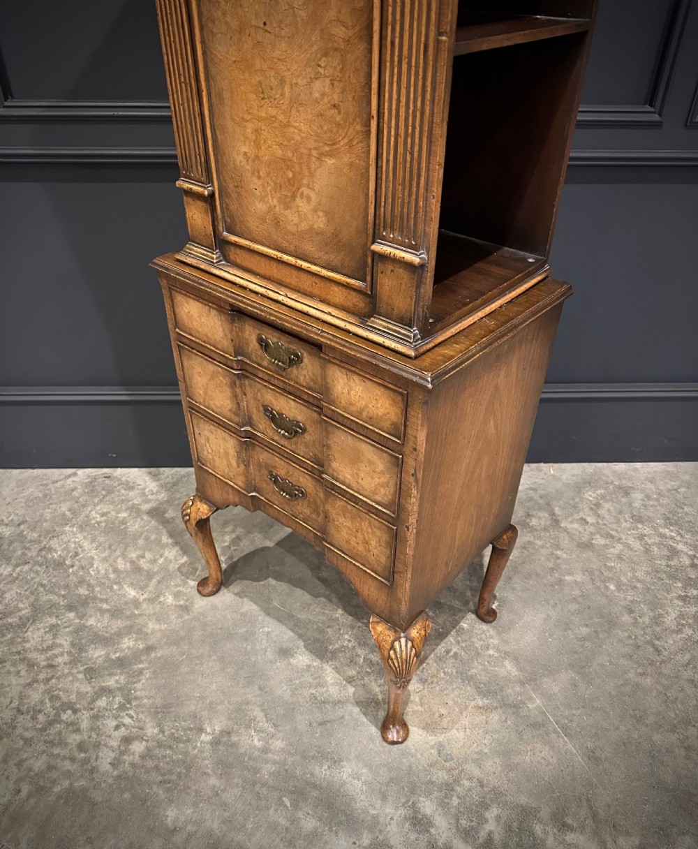 A tall, narrow Queen Anne style walnut bookcase featuring an arched burr walnut front panel, open side shelving, three drawers with brass handles, and cabriole legs with carved shell knees. View VII