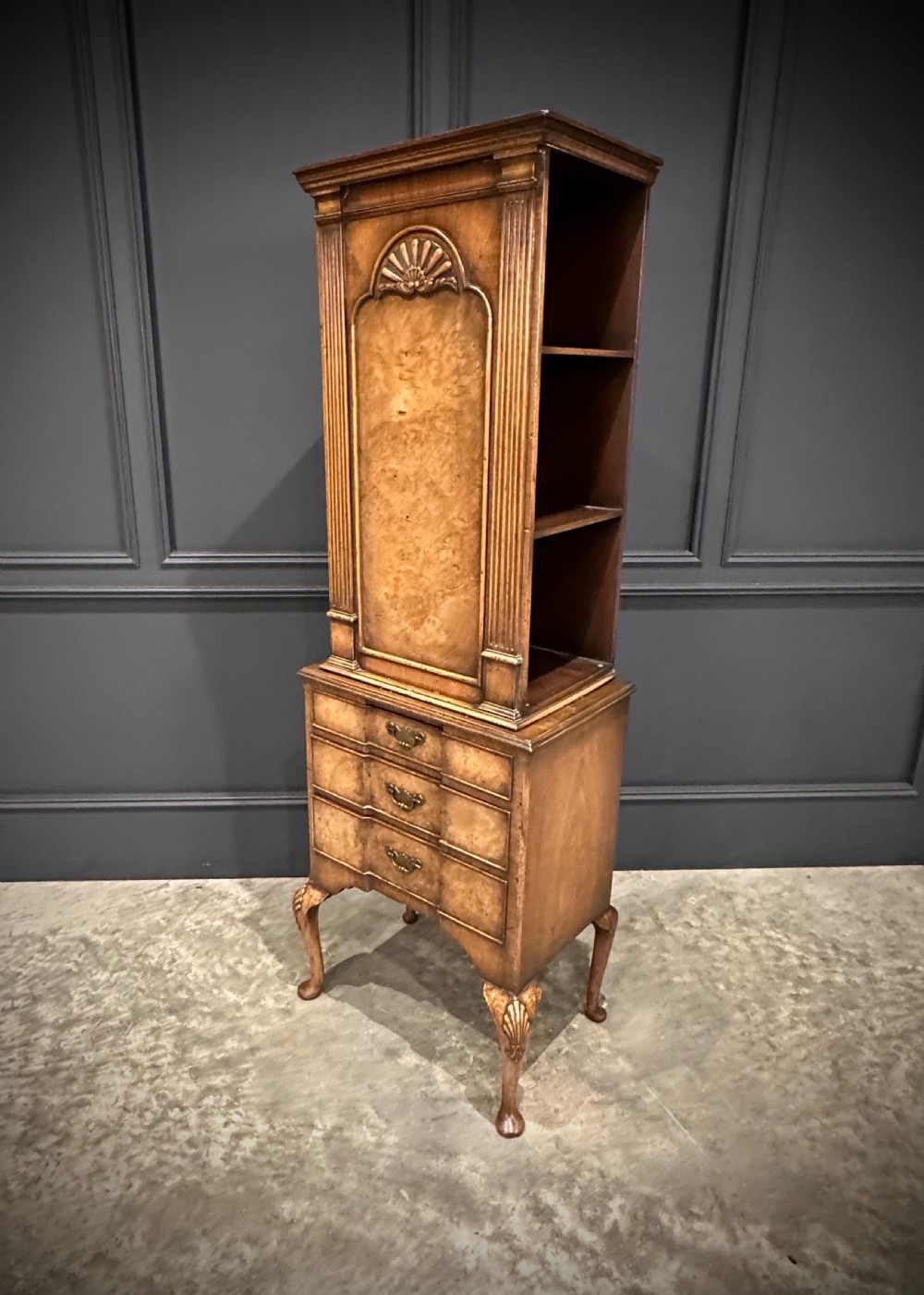 A tall, narrow Queen Anne style walnut bookcase featuring an arched burr walnut front panel, open side shelving, three drawers with brass handles, and cabriole legs with carved shell knees. View VIII